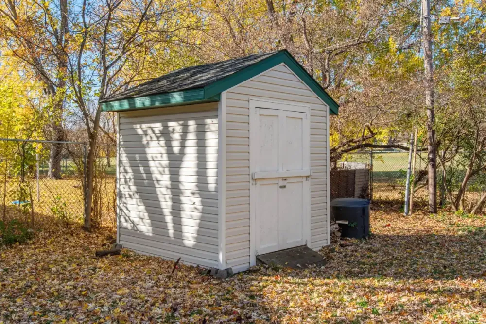 Vinyl-sided garden shed standing on a leaf-covered lawn behind a chain-link fence
